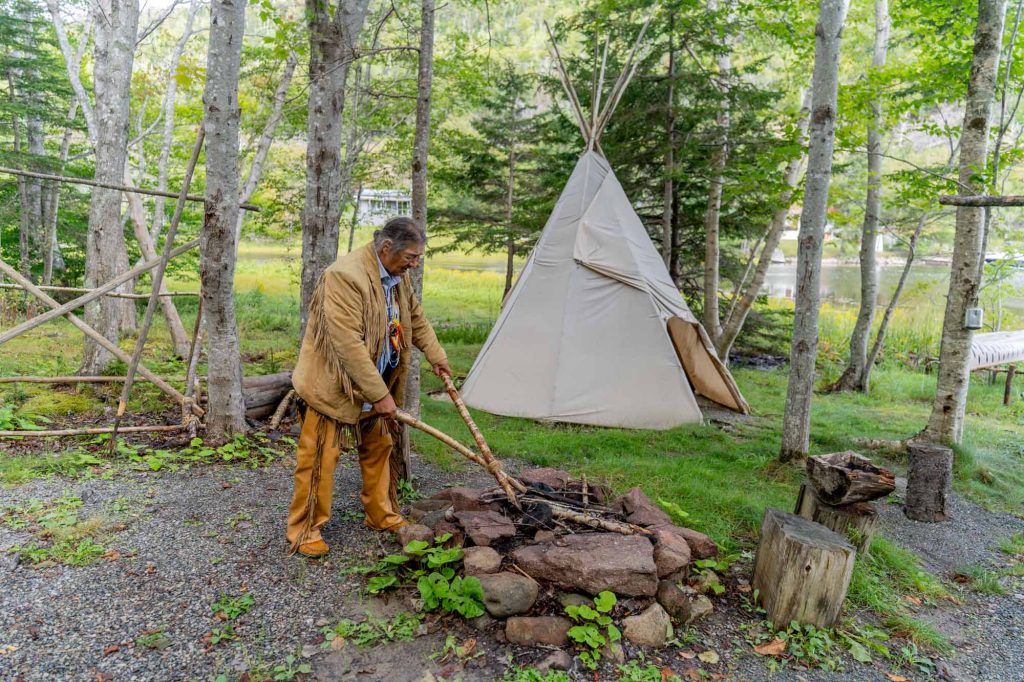 Indigenous man holding sticks over a fire with a tipi in the background