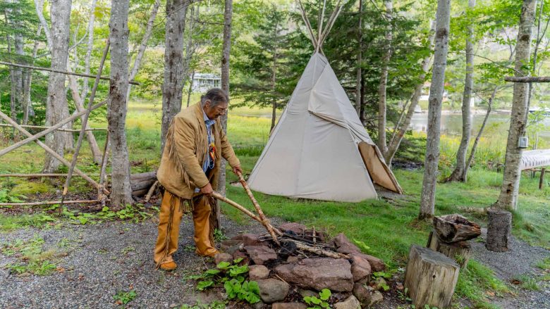 Indigenous man holding sticks over a fire with a tipi in the background