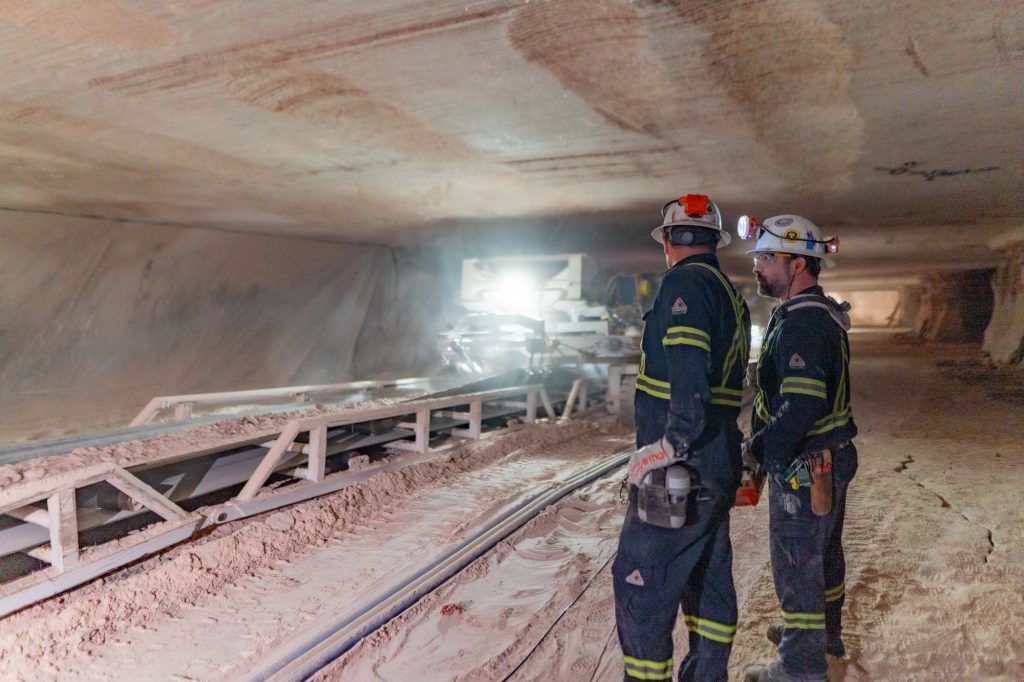 Two employees of Nutrien watch potash move on a conveyor belt in a potash mine.